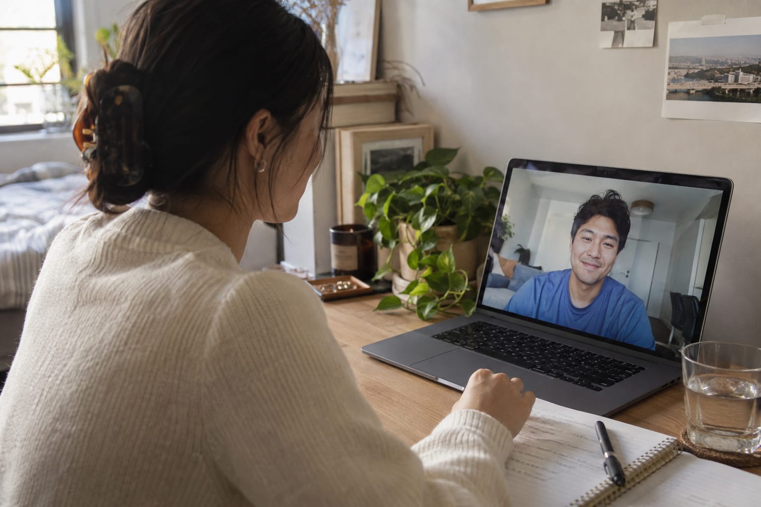 A woman at a desk talks with a smiling man on a laptop video call, with a notebook and plants nearby.