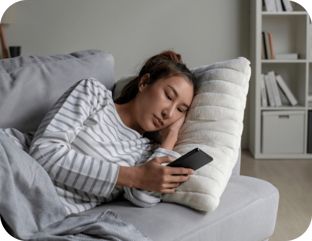 A woman in a striped shirt lies on a grey couch, resting her head on a white pillow while holding and looking at her smartphone, perhaps taking a moment for a Limerence Test. Shelves with books and decorative items are in the background.