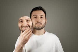 A man in a white shirt holds a smiling mask in front of his face, revealing a neutral expression underneath—a striking depiction of ADHD masking and the effort to hide true feelings.