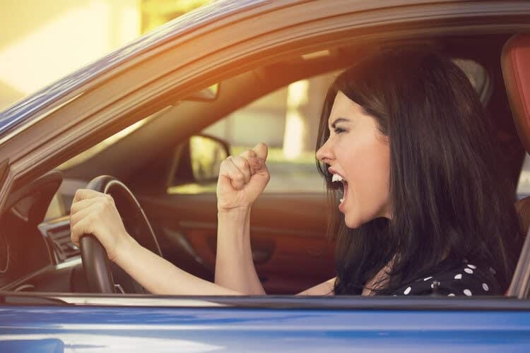 A woman in a blue car shouts and raises her fist while gripping the steering wheel with her other hand, clearly in need 