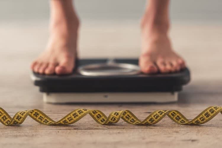 Bare feet standing on a weighing scale with a yellow measuring tape lying in front of the scale on the floor, perhaps ju