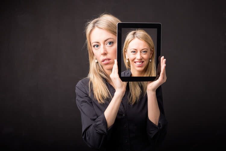 A woman holds a tablet in front of her face. The tablet displays an image of her smiling, while her actual expression is