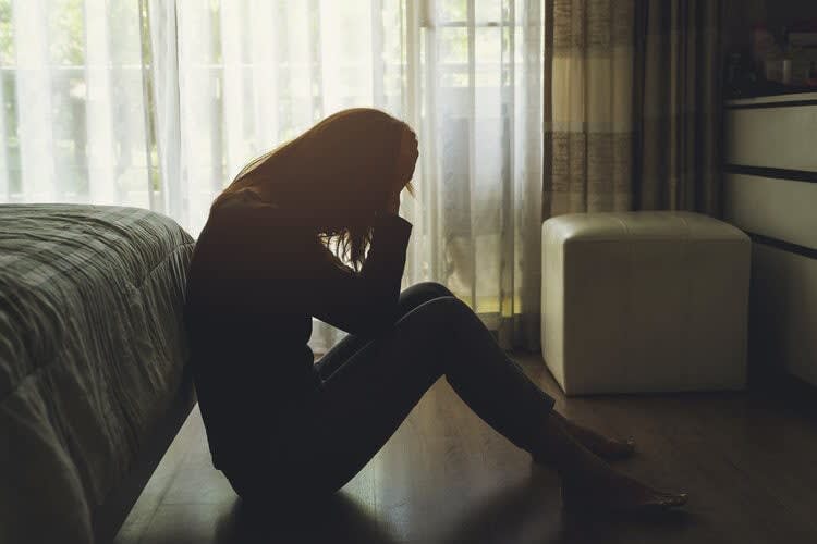 A silhouetted person sits on the floor beside a bed in a dimly lit room, cradling their head in both hands. Sunlight fil