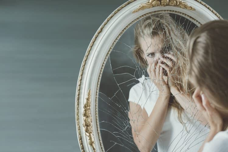 A woman wearing a white shirt stands in front of a cracked oval mirror, covering her mouth with her hands as she looks a