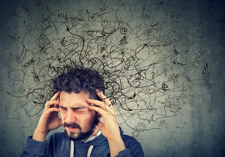 A man with a beard and closed eyes holds his temples while a chaotic swirl of abstract lines and symbols emanates from h
