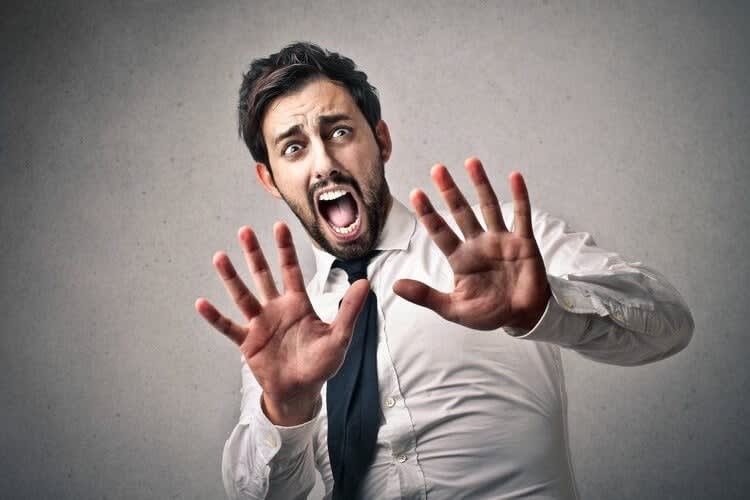 A man in a white shirt and tie is captured with a frightened expression, holding up both hands in a defensive gesture. I