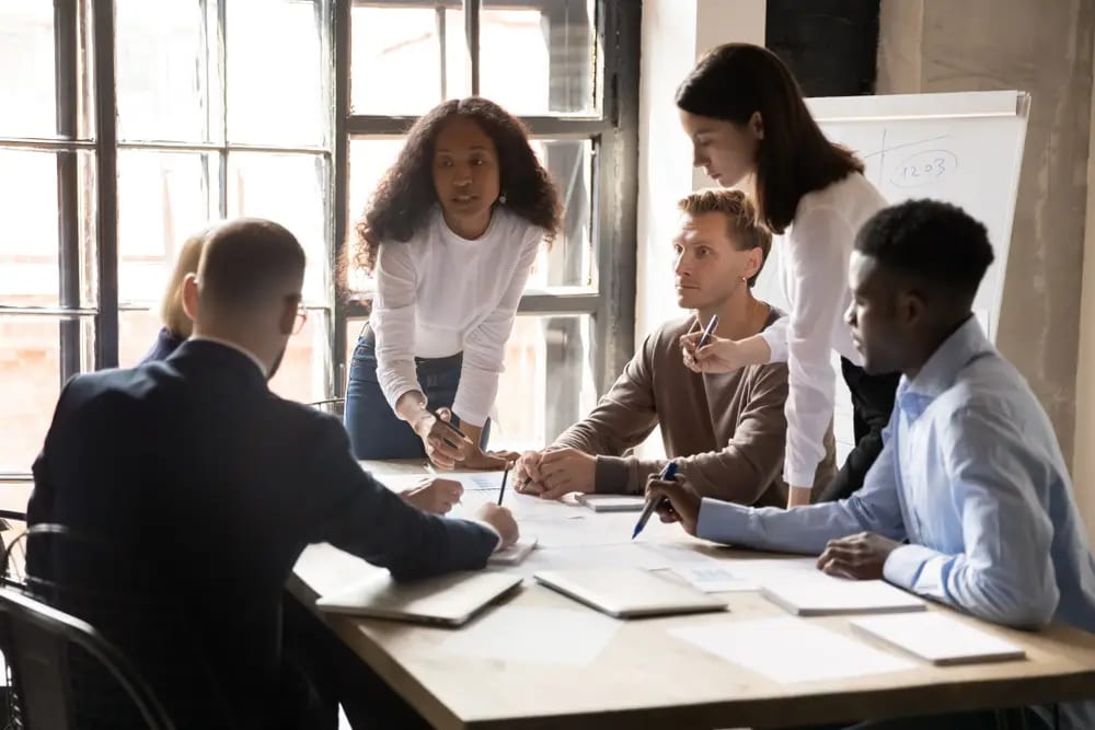 A diverse group of five people are gathered around a table in an office, engaging in a discussion and examining document