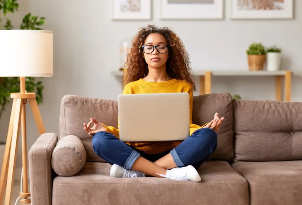 A person with curly hair and glasses sits cross-legged on a couch, balancing a laptop on their lap. With eyes closed and