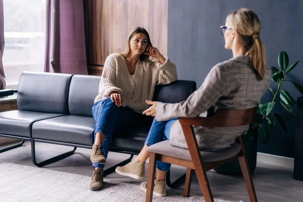 Two women are sitting in a living room having a conversation. One woman is on a sofa, the other on a chair. The room, re