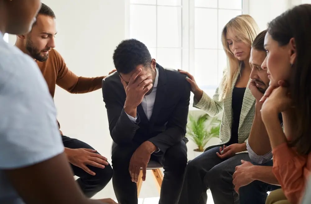 A group of six people sit in a circle, with some placing comforting hands on a person in the center who is covering thei