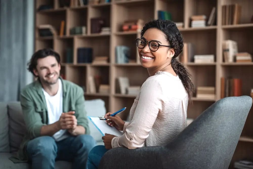 A woman with glasses takes notes on a clipboard while smiling and sitting in a chair. A man sits across from her on a co