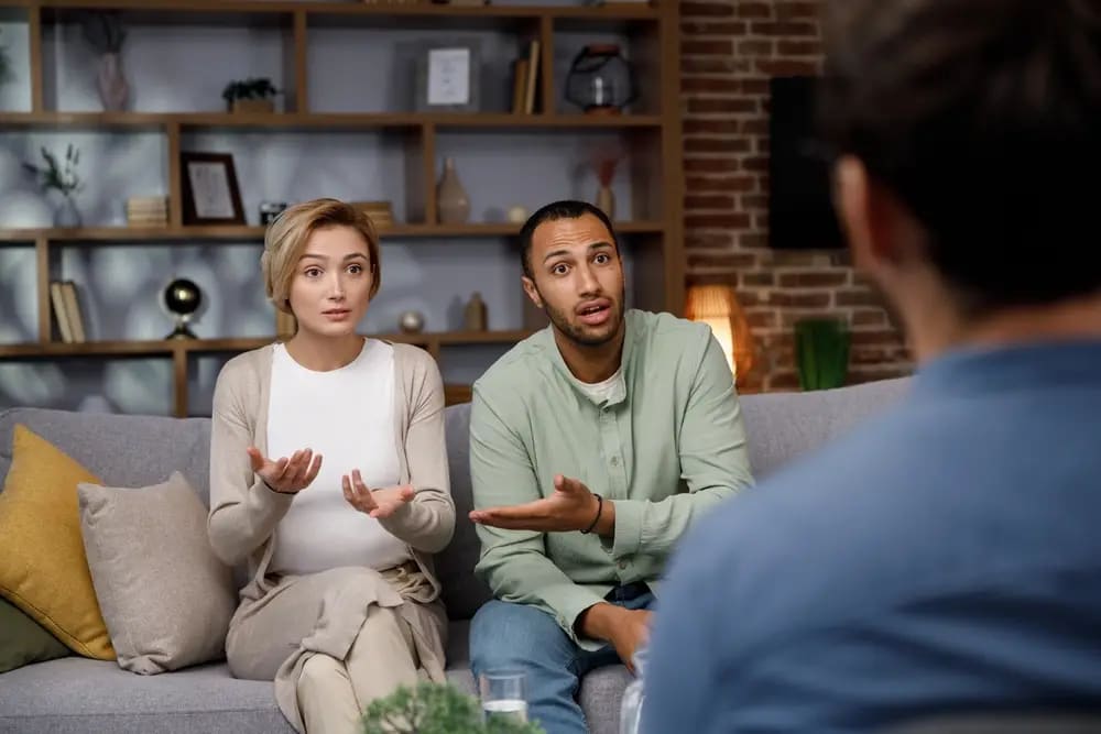 A woman and a man are sitting on a couch, gesturing and talking to a Roseville therapist opposite them. Shelves with dec