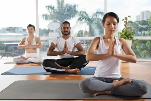 Three people sitting on yoga mats in a peaceful room, practicing meditation with hands in prayer position, eyes closed. 
