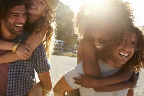 Four friends smiling and laughing, with two giving piggyback rides at a sunny beach, embody the joy often rediscovered t
