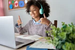 Young girl with headphones using a laptop and giving a thumbs up at a desk with books and papers, during an online therapy session. Choosing your therapy online vs in person