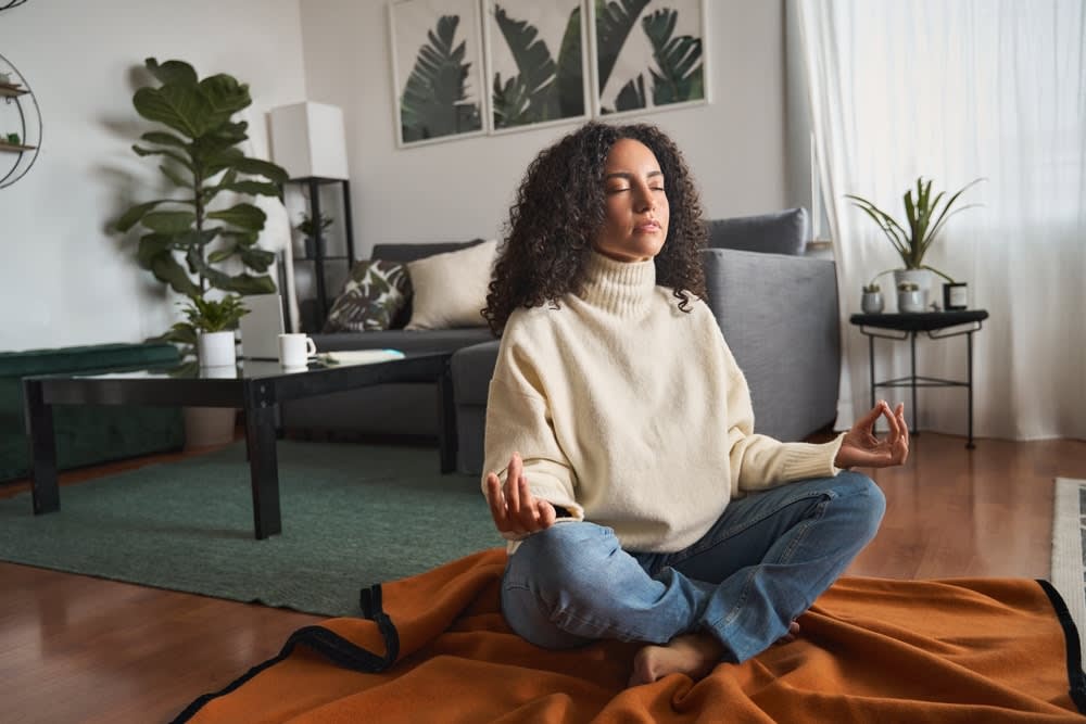 A somatic therapist SF with curly hair, wearing a white sweater and jeans, sits cross-legged on a blanket in a tranquil living room practicing meditation. The background includes serene plants, a cozy coffee table, and an inviting couch.