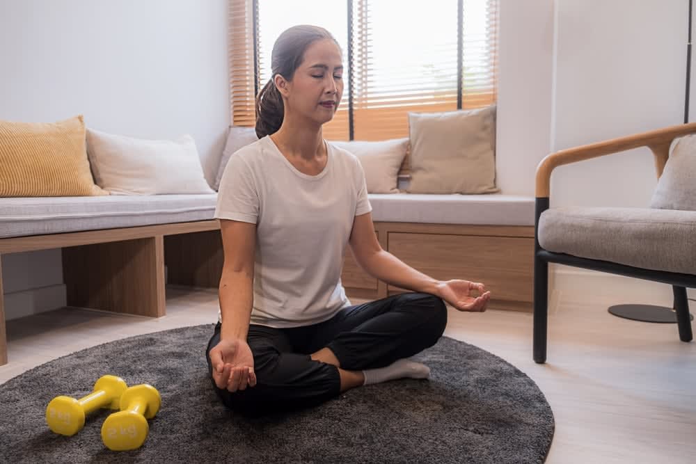 A person sits cross-legged on a rug in a living room, meditating. Yellow dumbbells are on the floor nearby, perhaps indicating the holistic approach of somatic psychotherapists in San Francisco who blend mindfulness with physical wellness.