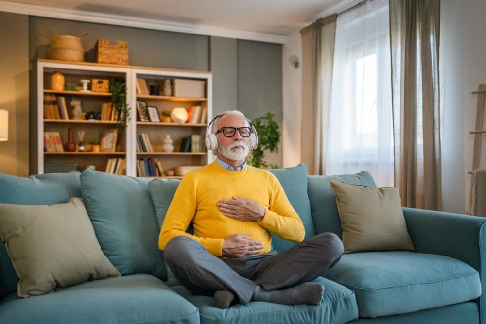 An elderly man with white hair and beard, wearing a yellow sweater and glasses, sits cross-legged on a blue sofa in the living room with bookshelves and decor, meditating with one hand on his chest—a practice often encouraged by somatic therapists in San Francisco.