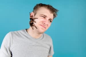 A man in a grey t-shirt grimaces as a large tarantula crawls on the side of his face against a blue background, reflecting the profound journey of trauma and healing.