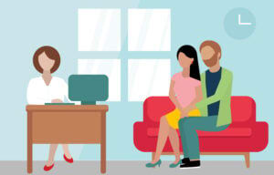 A couple sits on a red couch holding hands during a consultation with a premarital counselor seated at a desk in an SF therapy office, embodying the steps outlined in how to choose the right premarital counselors in San Francisco.