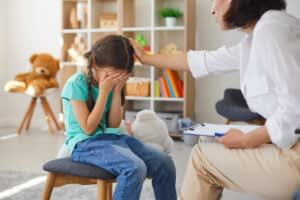 A San Francisco PE therapist comforts a crying girl in a prolonged exposure therapy session in SF. The girl covers her face with her hands while the woman, an ERP therapist, holding a clipboard, places a hand on her head. Toys are visible in the background.