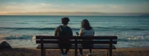 Two people are sitting on a bench at the beach, facing the ocean under a clear sky with gentle waves rolling in. One person is looking at a smartphone while they both enjoy the scenic view, sharing a moment of harmony amidst nature's beauty.