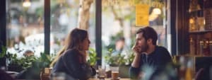 Two people sit across from each other at a café, engaged in conversation. Beverages are on the table, and the background shows a window with plants and outdoor scenery. Their interaction highlights the principles of Effective Listening for Couples, fostering deeper connection and understanding.