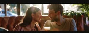 A man and woman sit at a sunlit table in a restaurant, smiling and looking at each other. A blurred drink is in the foreground as they engage in crafting healthy emotional dialogue, fostering a deeper connection in their marriage.