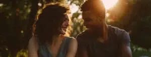 Two people sit closely together outdoors, smiling at each other with sunlight filtering through trees in the background, their non-verbal communication speaking volumes about their deep marital bond.