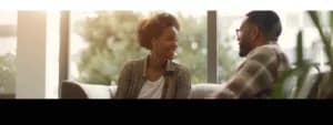 A woman and a man sitting on a couch, smiling and talking to each other, with a window and greenery in the background. Their conversation flows effortlessly, thanks to better communication strategies learned from couples therapy.