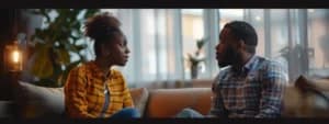 Two people sit on a couch having a serious conversation in a well-lit room with large windows and plants in the background, the scene reminiscent of couples counseling in San Francisco.
