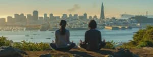 Two people sit cross-legged and meditate on a hill overlooking San Francisco Bay with the city skyline, including the Transamerica Pyramid, in the background.