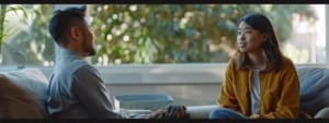 Two people sit on a sofa engaging in a conversation, with blurred greenery visible through the large window behind them. The scene captures the essence of a cozy San Francisco apartment as they discuss their thoughts with a CBT therapist.