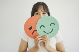 A woman holds a sad red face and a happy green face sign in front of her, partially covering her own face against a plain background, symbolizing the struggles of ADHD masking and revealing true emotions.
