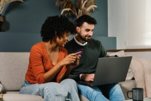 A woman holds a credit card while sitting next to a man using a laptop on a couch; both appear focused on the screen, highlighting the importance of managing finances in relationship dynamics.