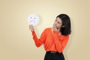 A woman in an orange sweater holds a sad face sign and touches her mouth, illustrating the struggles of ADHD masking, standing against a plain beige background.