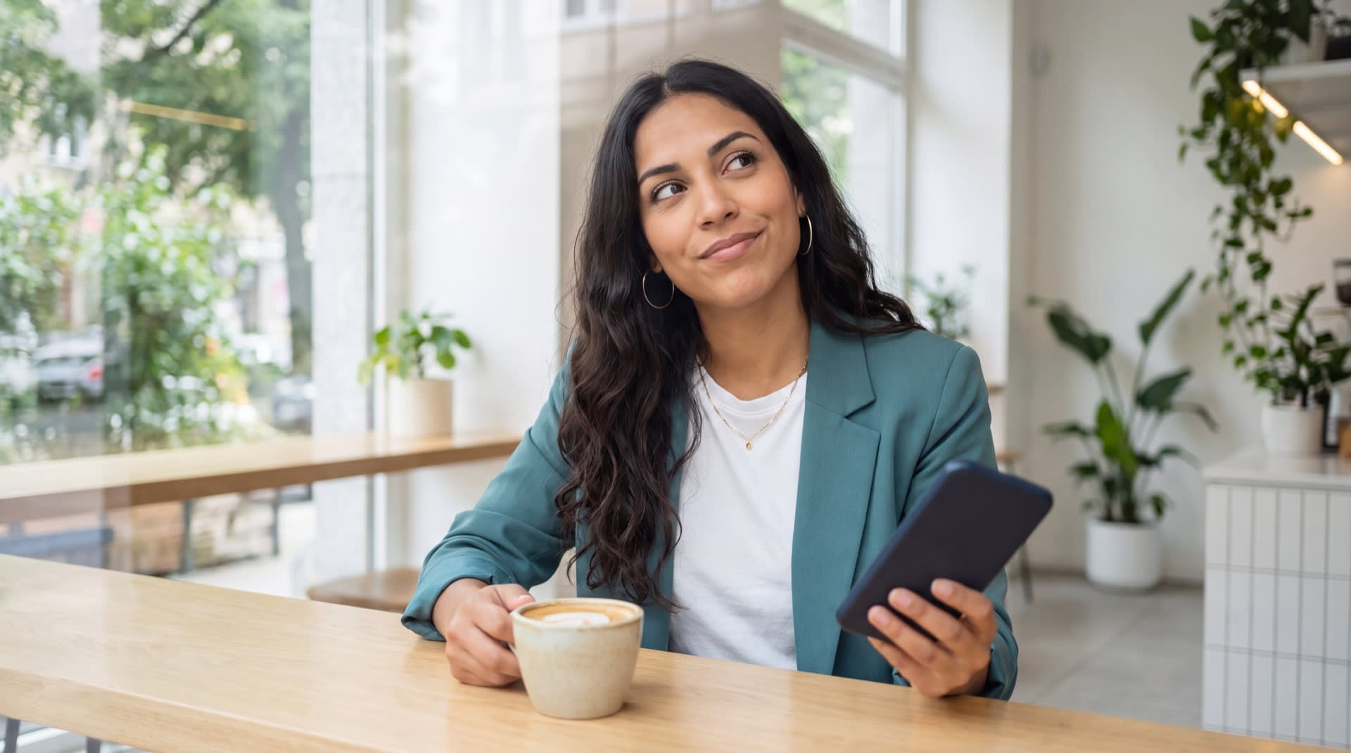 A woman sitting at a bright cafe window with her phone, pausing thoughtfully before responding to a message.