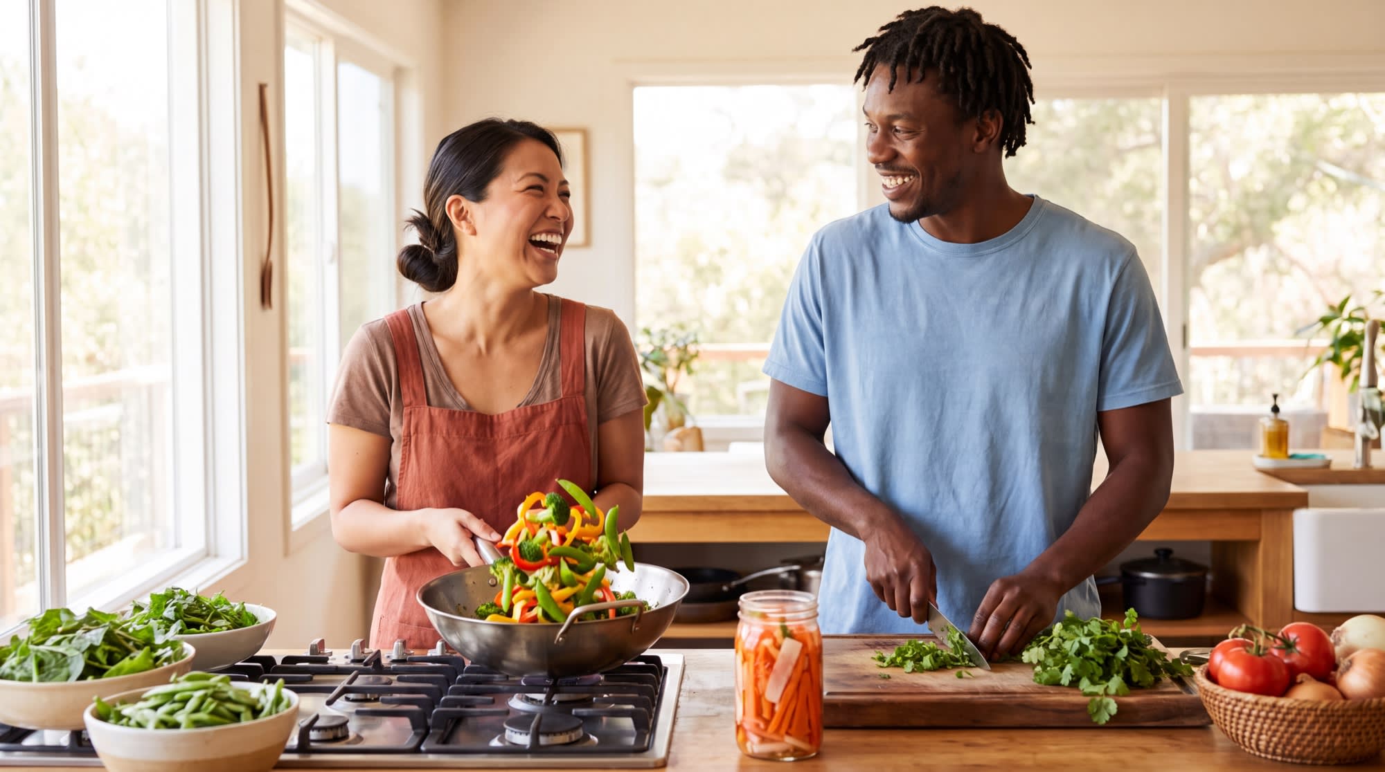 Two friends laughing while cooking a colorful vegetable stir-fry together in a bright kitchen.