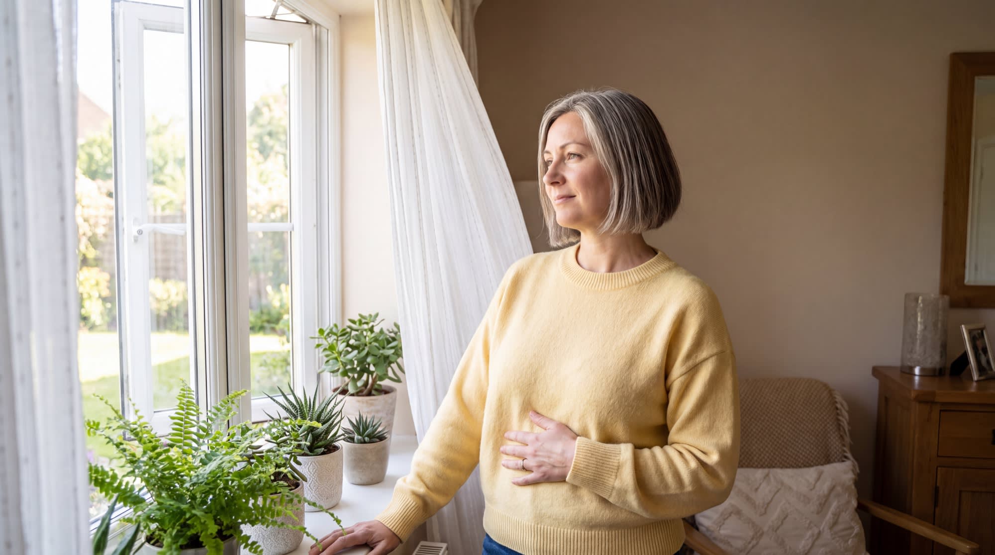 A woman standing at an open window with one hand on her belly, taking a slow deep breath in the morning light.