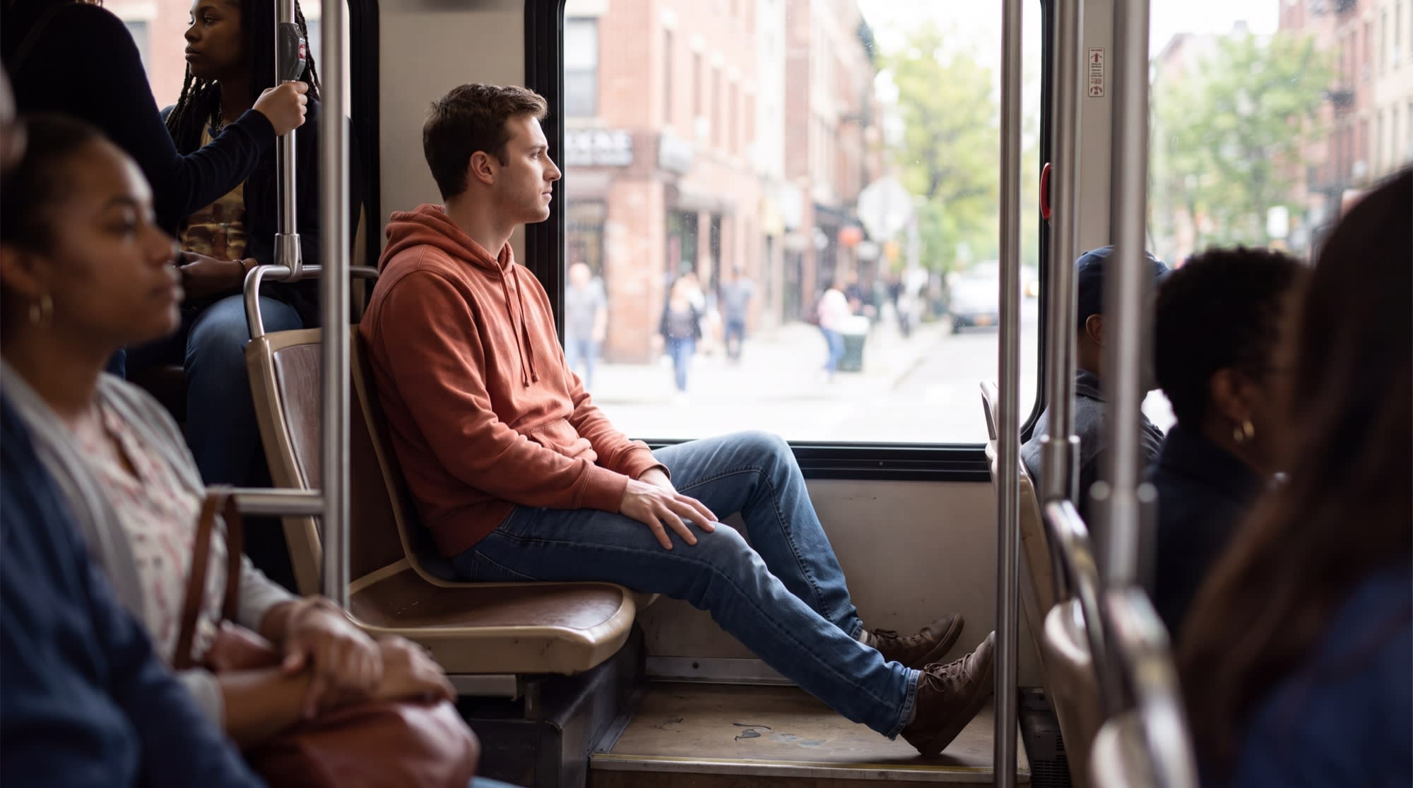 A man sitting by the window on a city bus with both feet flat on the floor, looking out calmly.