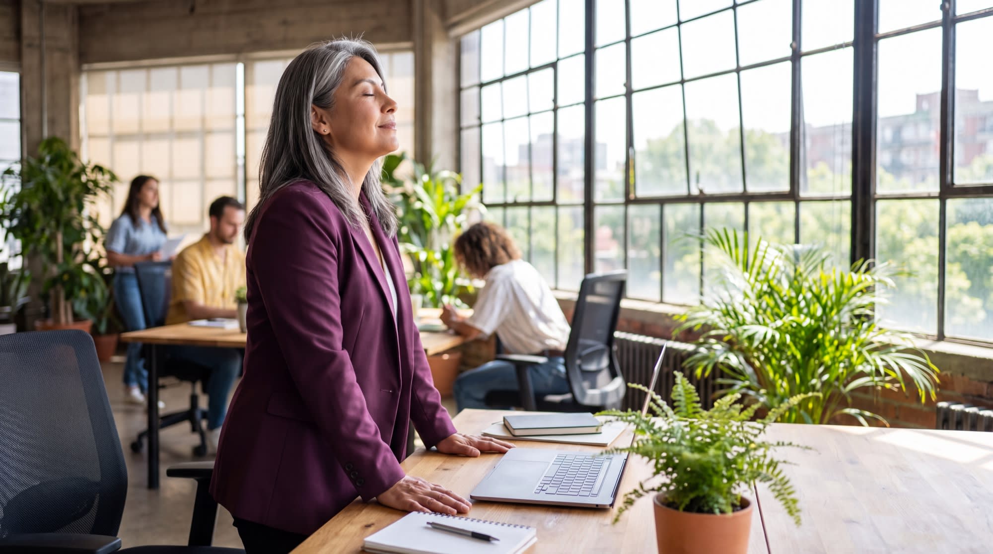 A woman pausing at her desk in a bright office, palms flat on the surface and eyes closed, taking a slow breath.