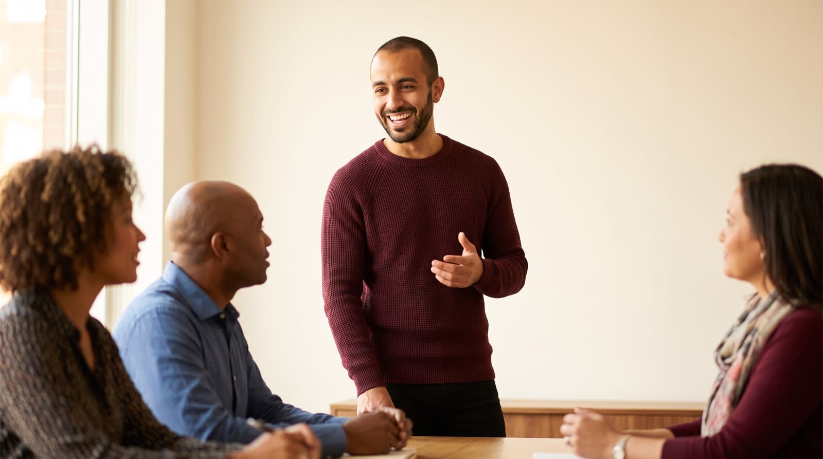 A man giving a relaxed, confident presentation to a small group of colleagues in a bright meeting room.