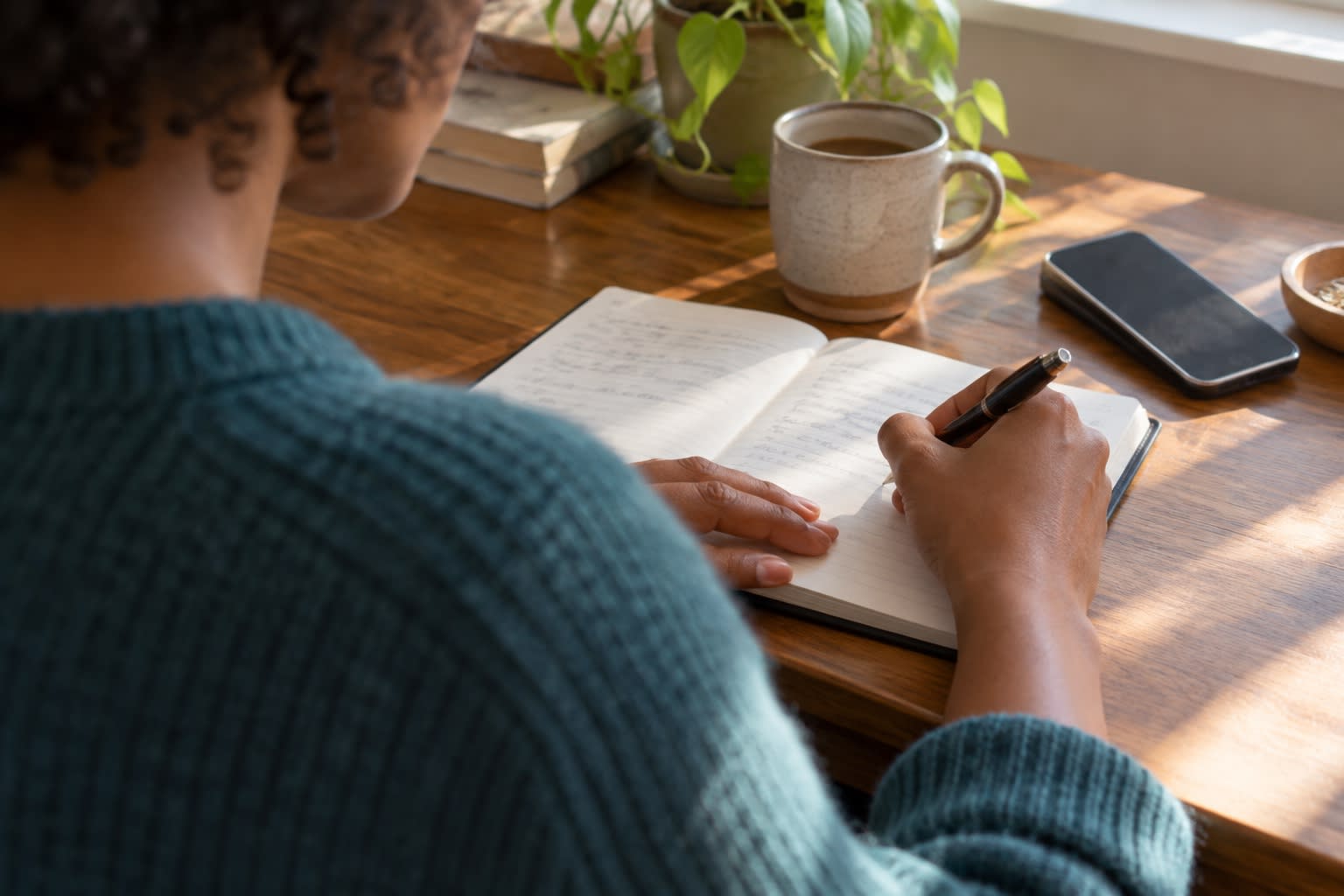 A person writes in a notebook at a sunlit wooden desk.