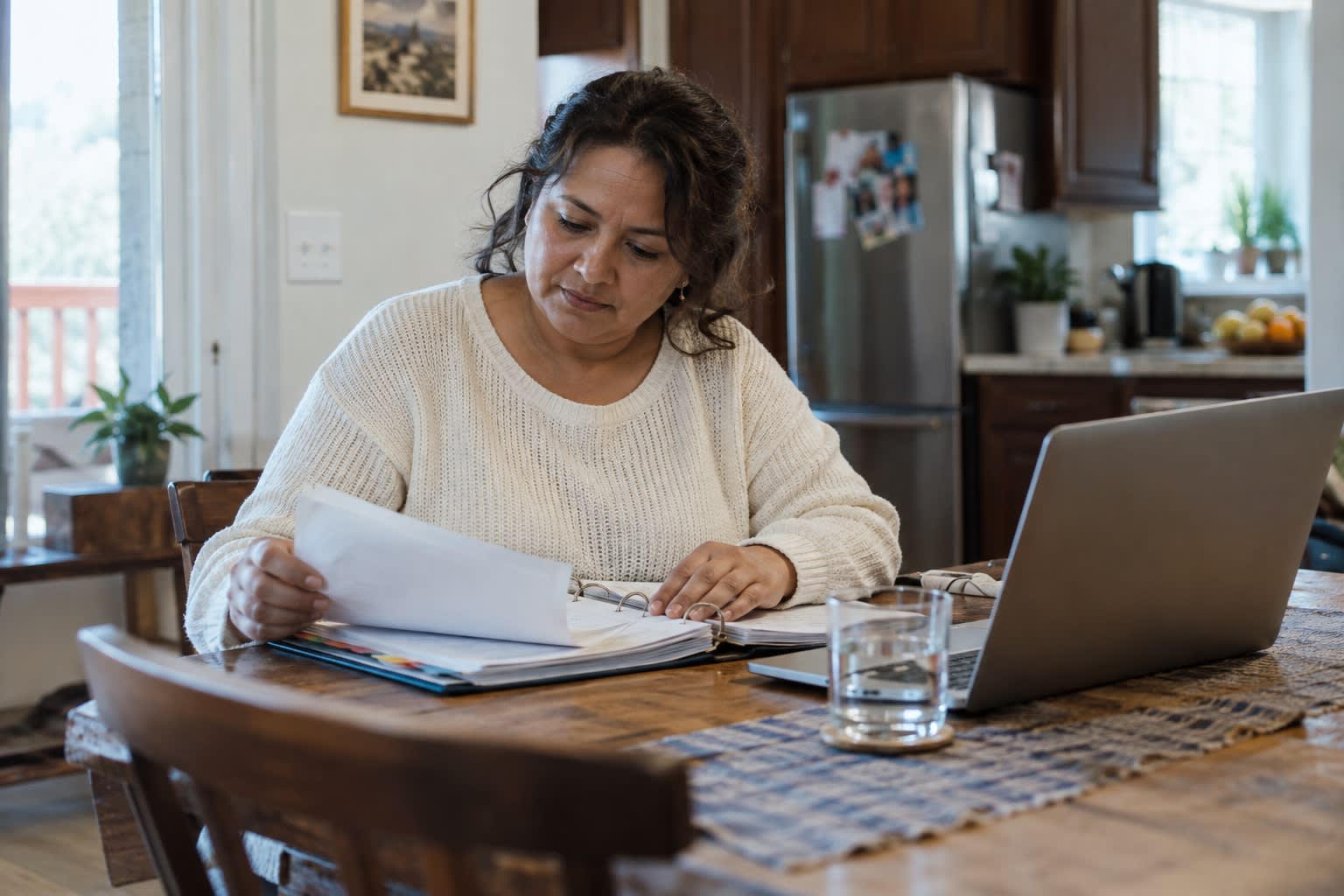 A woman reviews papers in a binder at her kitchen table next to a laptop, organizing documents for an ADHD disability application.