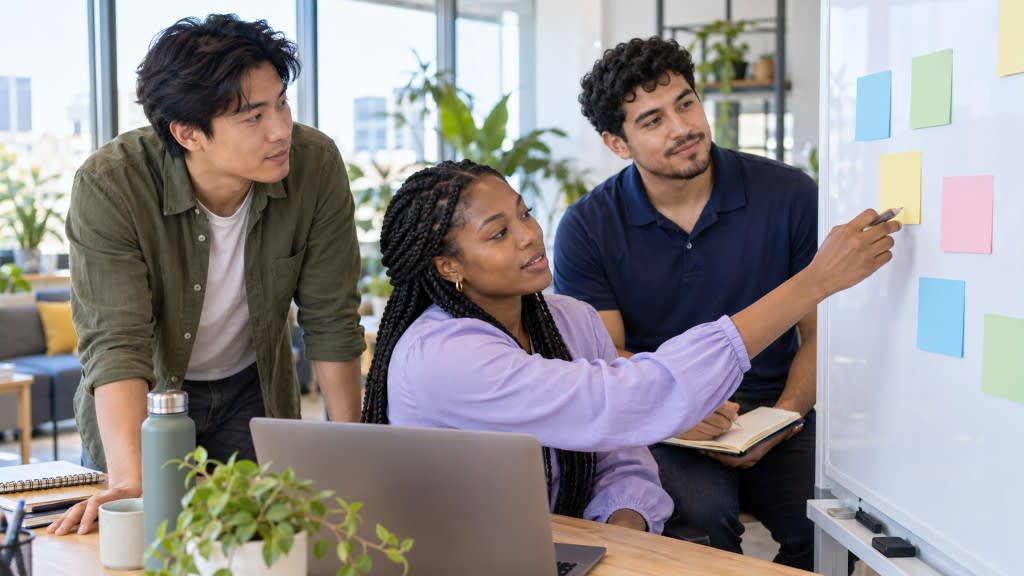 A small team of three colleagues is gathered around a whiteboard in a bright office room, engaged in a team meeting focused on setting SMART goals for their professional development and career success. They are discussing specific plans to achieve their personal and professional goals, emphasizing the importance of clear direction and measurable progress.