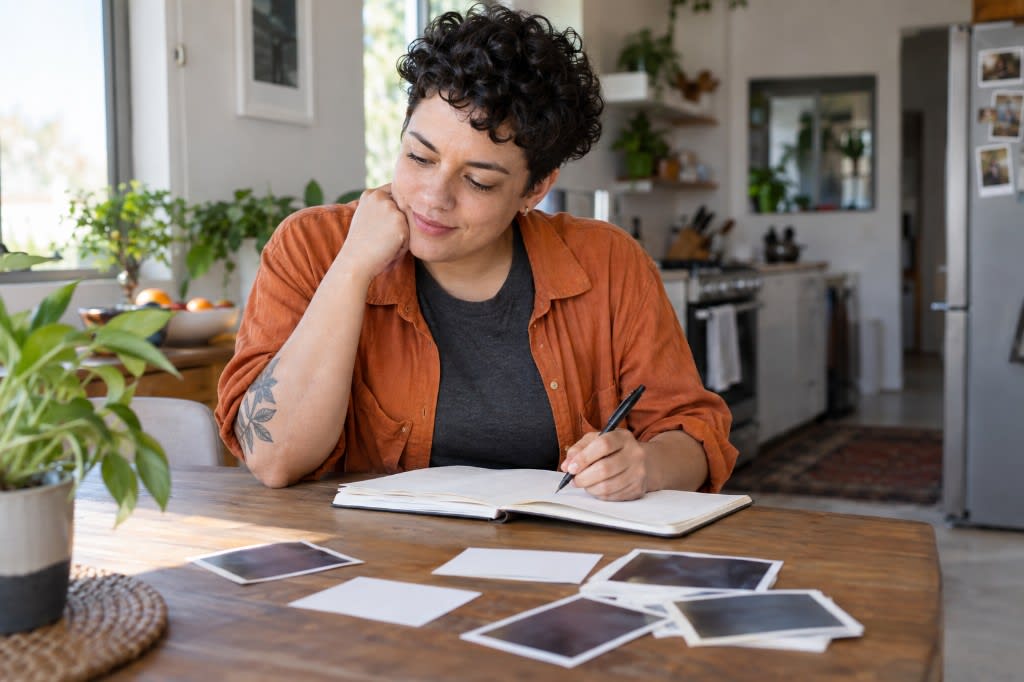 Someone journaling at a kitchen table with photographs spread out, tracing the pattern in past relationships