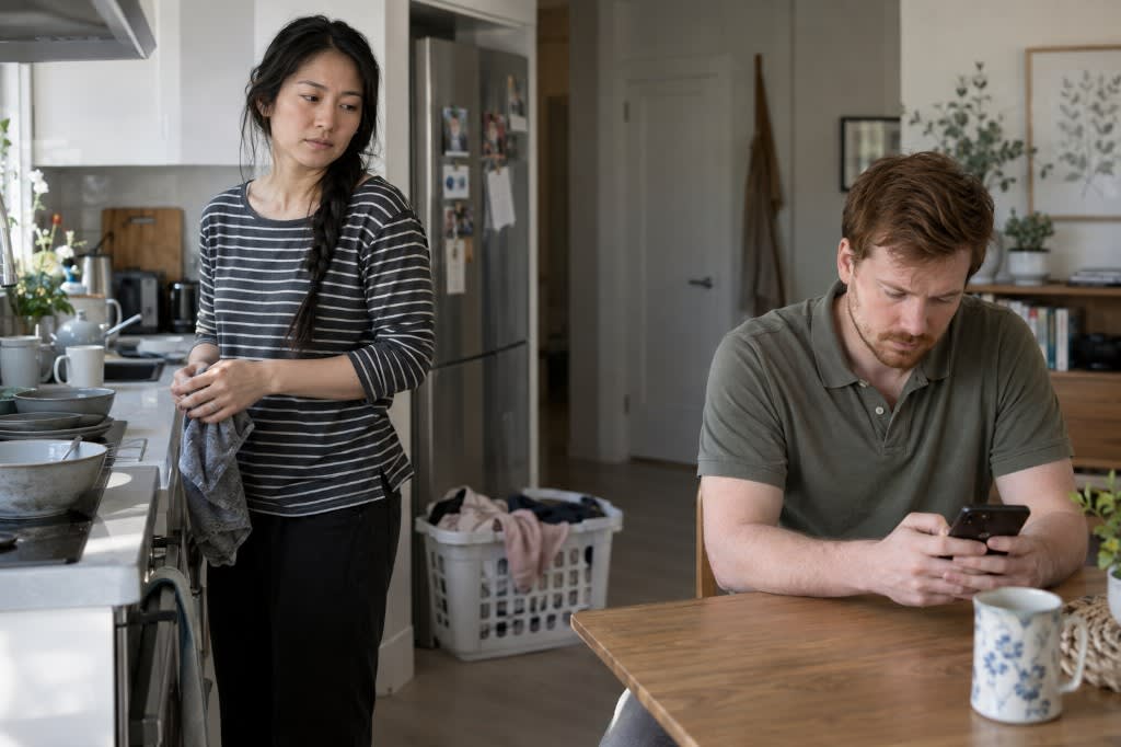 A woman holding a dish towel in the kitchen looking over at her partner scrolling his phone at the table beside a full laundry basket