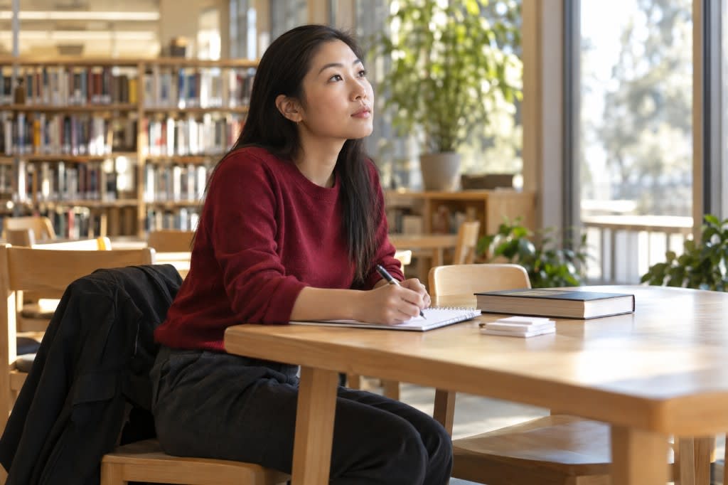 A student in a library looking up from her notebook, pen in hand, recalling material from memory before checking the closed book beside her