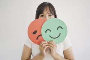 A woman holds a sad red face and a happy green face sign in front of her, partially covering her own face against a plain background, symbolizing the struggles of ADHD masking and revealing true emotions.
