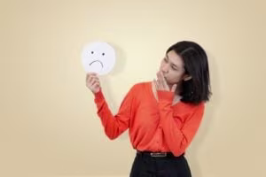 A woman in an orange sweater holds a sad face sign and touches her mouth, illustrating the struggles of ADHD masking, standing against a plain beige background.